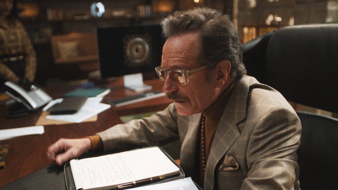 Bryan Cranston sitting at a desk with papers and a clipboard, wearing a suit and glasses. Two women, one pointing and wearing a sleeveless top with bracelets, the other in a checkered blazer and tie, sit at a table. A man in a brown suit stands in a room with large windows and mountains visible. Another man in a green shirt with headphones stands near a woman in a striped shirt, both in a warmly lit room with a painting on the wall.