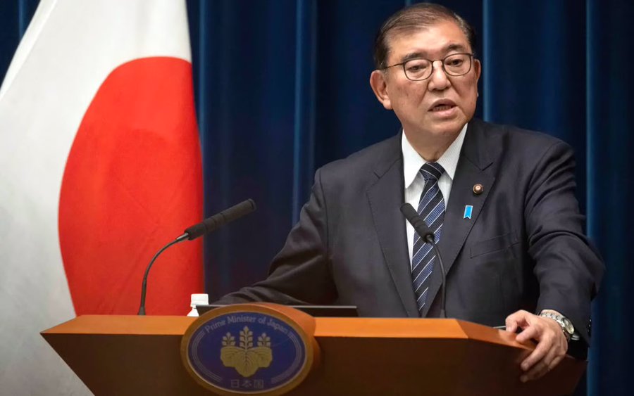 Shigeru Ishiba stands at a wooden podium with a microphone. He wears a dark suit, tie, and glasses. The Japanese flag is visible to his left, and a blue curtain forms the background.