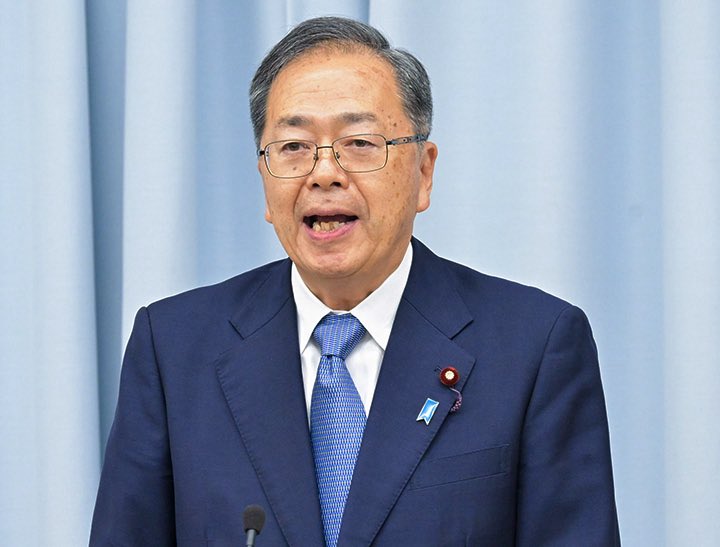 A man wearing a dark blue suit, white shirt, blue tie, and a pin on his lapel, standing in front of a light blue curtain. A microphone is visible in front of him.