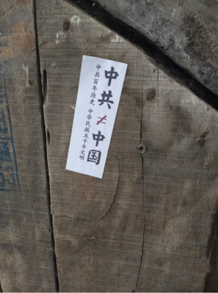木质表面，上面贴有白色的矩形贴纸，上面有黑色的中文文字。文字垂直对齐写着“中共共产党 伟大光荣正确 中华全国”。木材有可见的纹理和钉子或螺丝头。