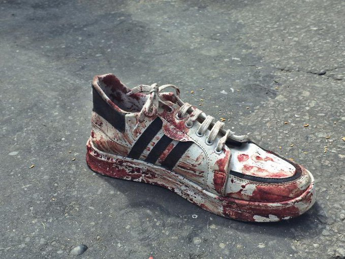 A single white sneaker with black stripes, resembling an Adidas design, lies on a cracked asphalt surface. The sneaker is heavily stained with red paint or blood, covering much of its surface, including the laces and sole. The background shows a rough, gray road with scattered debris.
