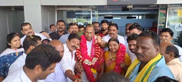 A group of people gathered at an airport arrival gate, some wearing white shirts and others in traditional attire with garlands. Petals are scattered on the ground, and a sign reads "ARRIVAL" in English and Telugu. Several individuals hold flowers and garlands, welcoming others.