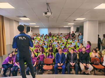 A large group of students wearing purple shirts stands in rows in a classroom, smiling at the camera. An astronaut in a blue and black suit with a Türkiye Uzay Ajansı patch holds a microphone on a stage, addressing the audience. Several adults in formal attire, including suits and dresses, stand among the students. A projector screen and tables are visible in the background.