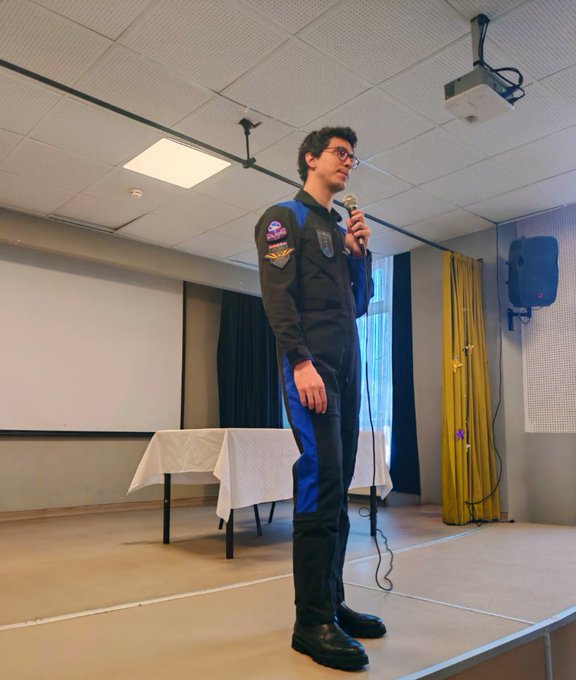 A large group of students wearing purple shirts stands in rows in a classroom, smiling at the camera. An astronaut in a blue and black suit with a Türkiye Uzay Ajansı patch holds a microphone on a stage, addressing the audience. Several adults in formal attire, including suits and dresses, stand among the students. A projector screen and tables are visible in the background.