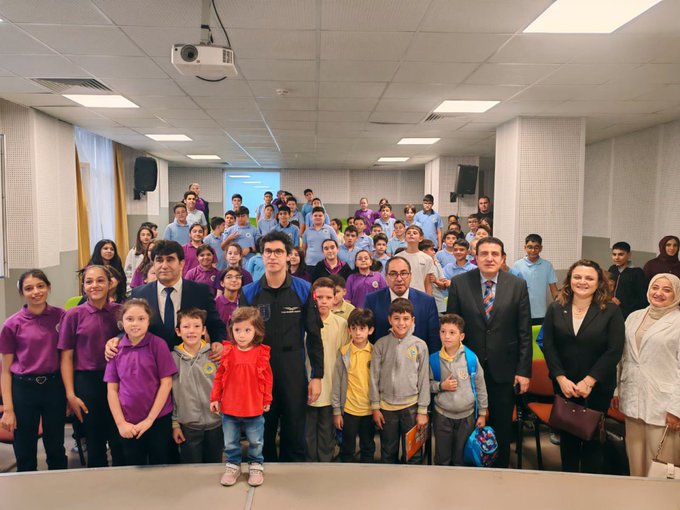 A large group of students wearing purple shirts stands in rows in a classroom, smiling at the camera. An astronaut in a blue and black suit with a Türkiye Uzay Ajansı patch holds a microphone on a stage, addressing the audience. Several adults in formal attire, including suits and dresses, stand among the students. A projector screen and tables are visible in the background.