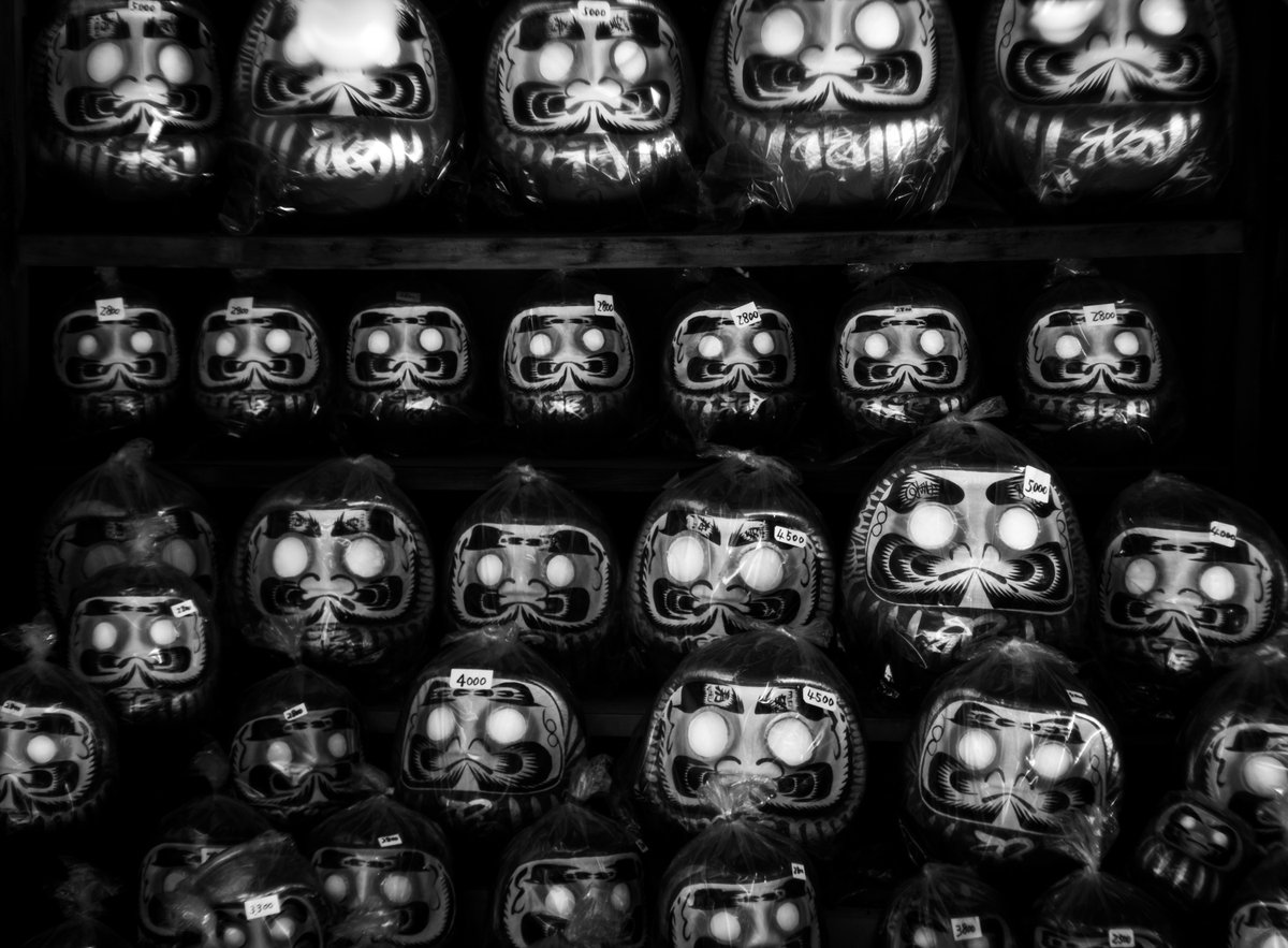 maxakisan's tweet photo. 【千の目力(Daruma’s Silent Pressure)】
薄暗い店先にぎょろっとこちらを見る目線。まだ白い目玉なのに、じっとこちらを見つめてくる。まるで「早く願いを書け！」と催促されているよう。 https://t.co/sOlSIORgDL