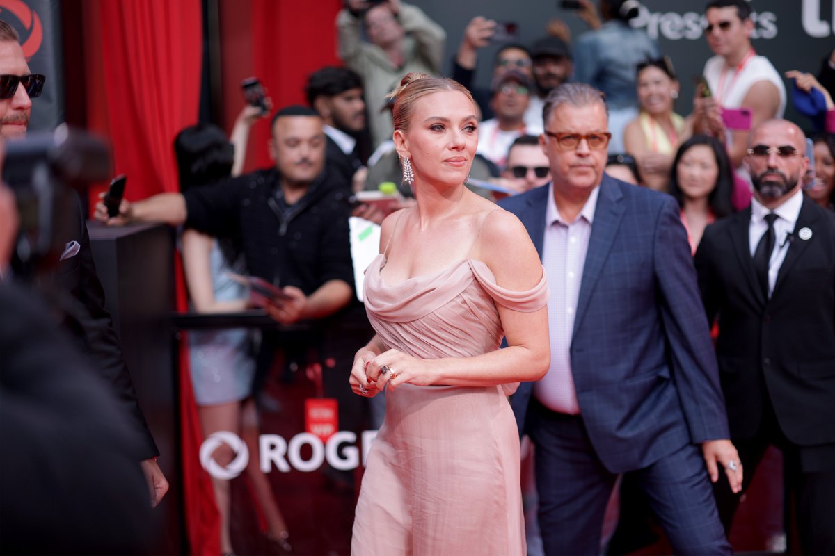 Scarlett Johansson wearing a pink off-shoulder dress on a red carpet. She is surrounded by photographers and security personnel. A man in a blue suit and glasses walks behind her.