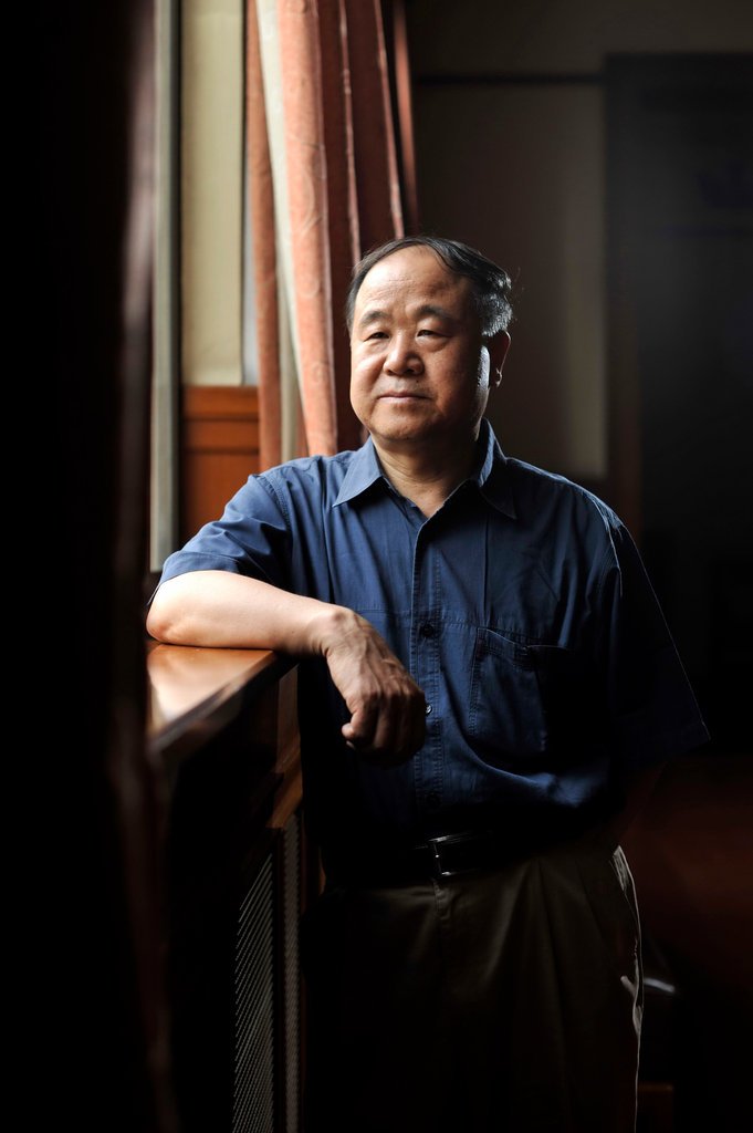 Mo Yan stands indoors near a window with curtains. He wears a blue short-sleeve shirt and beige pants, leaning casually on a wooden surface with one arm.