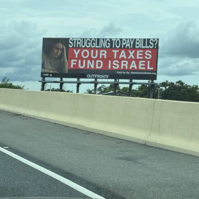 A billboard on a highway overpass with a woman looking down, her face partially visible. The billboard has bold text reading "STRUGGLING TO PAY BILLS? YOUR TAXES FUND ISRAEL" in white and red. The background shows a cloudy sky and trees, with the highway and guardrail visible below.