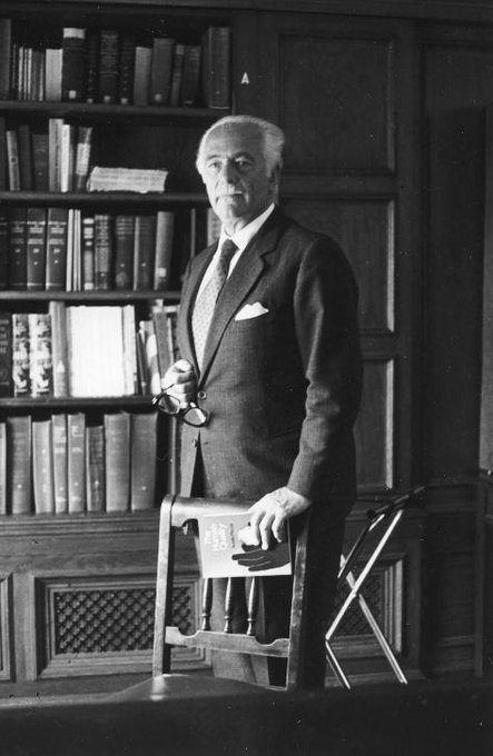 Aurelio Peccei stojí v obleku a kravatě, v jedné ruce drží brýle a v druhé papíry, stojí před policí plnou knih.