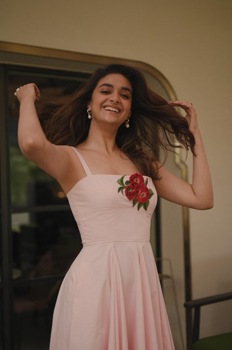 Keerthy Suresh wearing a sleeveless pink dress with red rose embroidery on the chest. Her long brown hair is loose, and she is smiling with her arms raised, holding her hair. She wears pearl earrings.