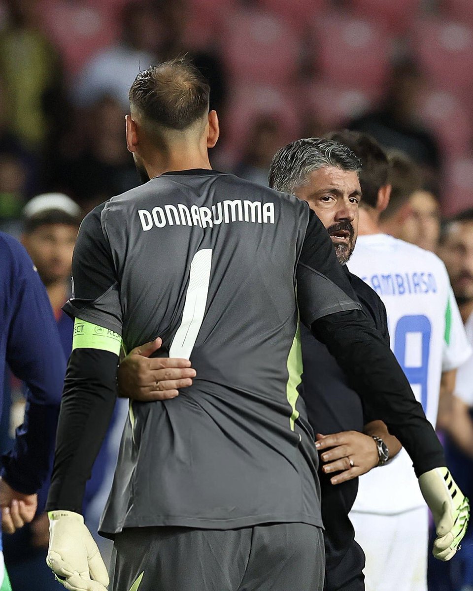 Gianluigi Donnarumma wearing a gray goalkeeper jersey with the name "DONNARUMMA" and number 1 on the back. He has gloves on his hands. A man with gray hair, wearing a dark jacket, stands behind him with arms around Donnarumma. Other players in jerseys are visible in the background, including one with "AMBROSIO" on it.