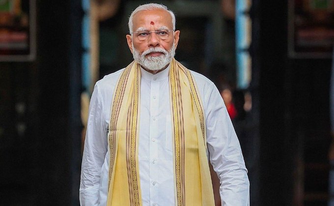 Narendra Modi wearing a yellow shawl and a colorful floral garland, walking outdoors with two men in black uniforms behind him. Pink architectural structures and a peacock design are visible in the background. In another image, Narendra Modi wearing a white shirt and yellow shawl, standing indoors with a dark background and blurred figures behind him.