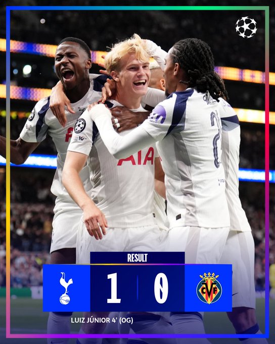 Three soccer players in white Tottenham jerseys with navy accents celebrating on a field. They are smiling and embracing, with the UEFA Champions League logo visible on their uniforms. A scoreboard shows Tottenham 1, VfB 0, with Luiz Junior credited for an own goal.