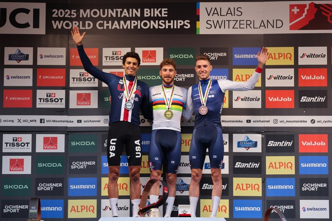 Victor Koretzky, Christopher Blevins, and Mathis Azzaro stand on a podium. Victor Koretzky wears a rainbow jersey and gold medal, Christopher Blevins wears a silver medal, and Mathis Azzaro wears a bronze medal. They raise their arms in celebration. The backdrop displays "2025 Mountain Bike World Championships Valais Switzerland" with logos for UCI, Tissot, Skoda, Shimano, and others.