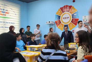 Mustafa Fatih Kacir stands in a room with children at tables, working with laptops and yellow robotics kits. The room has blue walls, a circular Deneyap logo, and a projector screen. Several children in casual clothing engage with the kits, while adults in formal attire observe.