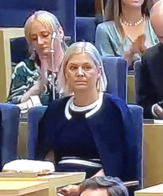 Magdalena Andersson sitting in a parliamentary setting, wearing a dark jacket with a white collar, surrounded by other individuals in blue seats, with microphones and papers visible on desks.