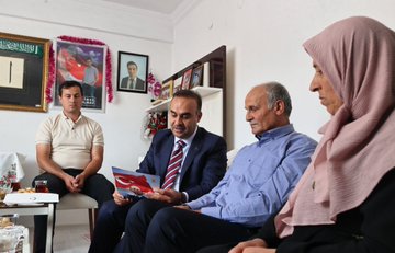 A man in a suit holding flowers stands at a doorway, greeting a woman in a headscarf. Inside a room, several people sit on couches, including a man in a suit holding a book with a red cover, another man in a blue shirt, and a woman in a headscarf. The room has framed photos, a Turkish flag, and floral decorations on shelves.