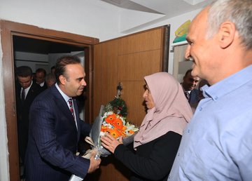 A man in a suit holding flowers stands at a doorway, greeting a woman in a headscarf. Inside a room, several people sit on couches, including a man in a suit holding a book with a red cover, another man in a blue shirt, and a woman in a headscarf. The room has framed photos, a Turkish flag, and floral decorations on shelves.