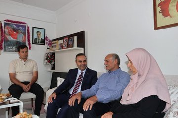 A man in a suit holding flowers stands at a doorway, greeting a woman in a headscarf. Inside a room, several people sit on couches, including a man in a suit holding a book with a red cover, another man in a blue shirt, and a woman in a headscarf. The room has framed photos, a Turkish flag, and floral decorations on shelves.