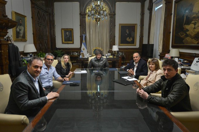 Javier Milei sits at the center of a large wooden table in an ornate room with chandeliers, portraits, and an Argentine flag. Six other individuals, including men and women in formal attire, sit around the table, some holding documents. The room features dark wood paneling, classic furniture, and large windows with curtains.