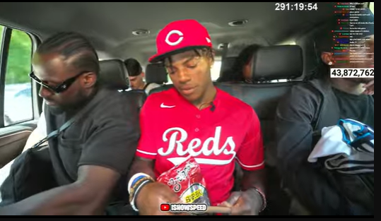 Darren Watkins Jr., wearing a red Cincinnati Reds jersey and matching cap, sits in a vehicle. He holds a bag of snacks and wears bracelets on his wrist. Other individuals are visible in the background, seated in the vehicle.