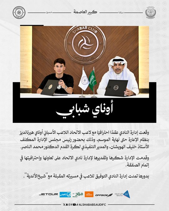 Two men sitting at a table with laptops. One man wears a black shirt, the other wears a white robe and headscarf. The Al Shabab Club logo is visible on a banner behind them. Saudi Arabian flags are on the table.