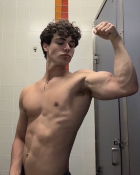 Zackary Arthur flexing his muscular arm in a gym, wearing a black fitted shirt and dark pants. His physique is well-defined, showing toned abs and biceps. The gym has exercise equipment and other people in the background. In another image, Zackary Arthur is shirtless, flexing his arm in a locker room with tiled walls and a metal door.
