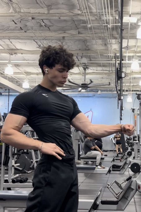 Zackary Arthur flexing his muscular arm in a gym, wearing a black fitted shirt and dark pants. His physique is well-defined, showing toned abs and biceps. The gym has exercise equipment and other people in the background. In another image, Zackary Arthur is shirtless, flexing his arm in a locker room with tiled walls and a metal door.
