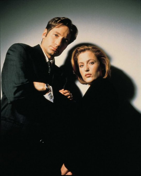 David Duchovny and Gillian Anderson standing close together. Both are dressed in dark suits. The background is a plain, light-colored wall with dramatic shadows.