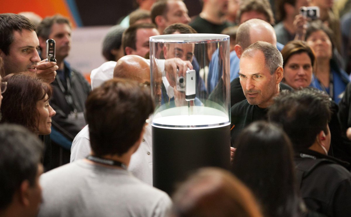 A crowd of people gathered around a clear cylindrical display case containing an iPhone. Steve Jobs stands nearby, looking at the device. Many individuals hold cameras or phones, capturing the moment.