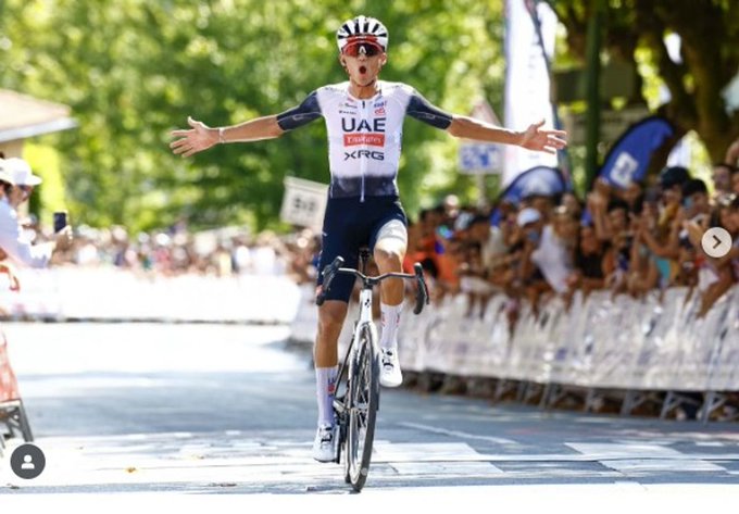 Isaac del Toro riding a bicycle, wearing a white and blue cycling jersey with "UAE" and "Team" text, a helmet, and sunglasses. His arms are raised in celebration as he crosses a finish line on a road lined with spectators and barriers.