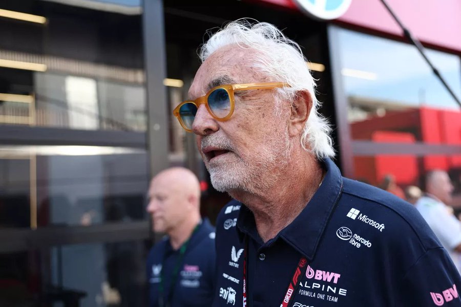 Flavio Briatore wearing a dark blue Alpine team shirt with logos including Microsoft, DSWT, and Alpine. He has white hair and is wearing yellow-tinted sunglasses. The background shows a building with glass windows and red elements, with another person in a similar team uniform visible.