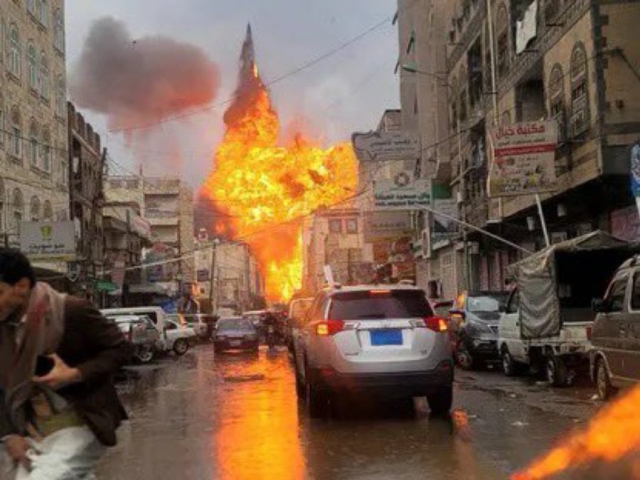 A street scene with a massive explosion in the background, flames and smoke rising into the sky. Cars are visible on the wet road, and buildings line the street with visible signs in Arabic. A person in a dark coat stands in the foreground, and debris or flames are near the bottom of the image.