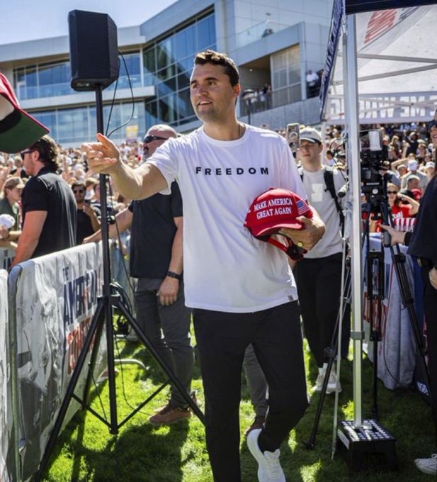A man wearing a white "FREEDOM" t-shirt and black pants, holding a red "Make America Great Again" hat, stands on grass at an outdoor event. He points forward with his right hand, surrounded by a crowd and tents. A speaker stand and banners with text are visible in the background.