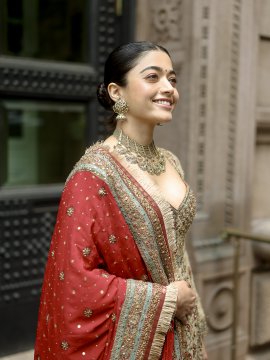 Rashmika Mandanna wearing a red and beige traditional outfit with intricate embroidery. She has a matching red dupatta draped over her shoulder. She wears elaborate gold jewelry, including a necklace and earrings. Her hair is styled in a neat bun.