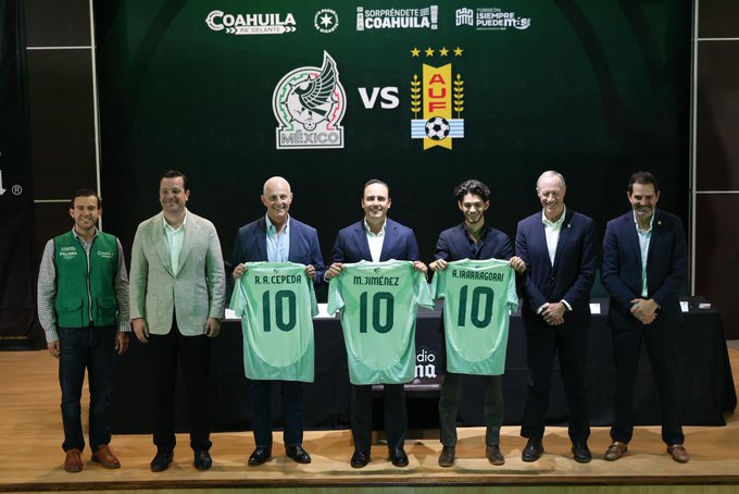 Seven men standing on a stage, holding green soccer jerseys with names R. Cárdenas, R. Jiménez, and R. Rodríguez, each with the number 10. A backdrop displays logos for Coahuila, the Mexico national team, and Uruguay, with text reading "MÉXICO VS URUGUAY." The setting includes a green background with additional text and symbols related to the event.