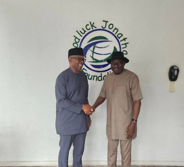 Peter Obi and Goodluck Jonathan shaking hands, both wearing traditional Nigerian attire. Peter Obi is in a gray outfit with a black cap, and Goodluck Jonathan is in a beige outfit with a black hat. A logo behind them reads "Goodluck Jonathan Foundation" with text and a circular design.
