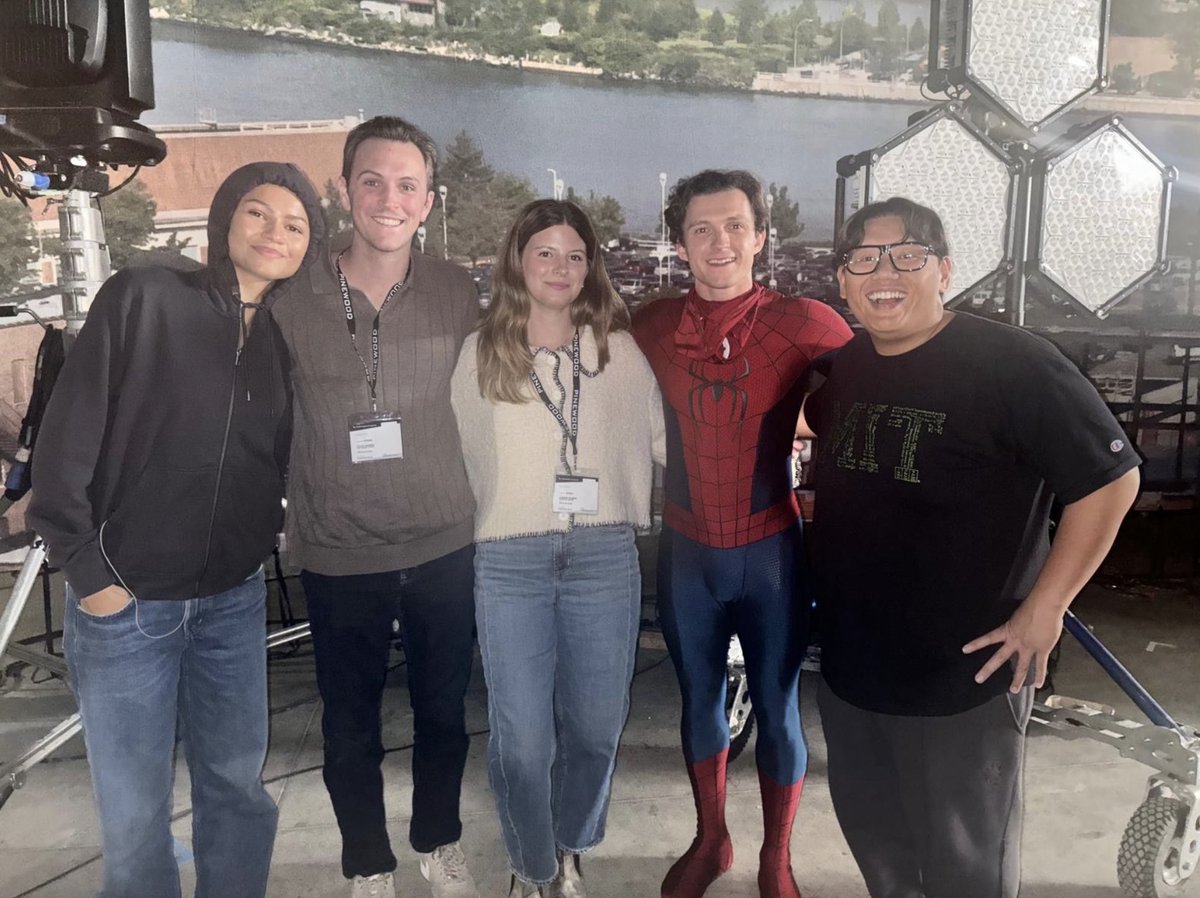 Tom Holland in a Spider-Man costume, Zendaya in a black hoodie, Jacob Batalon in a brown shirt, and two other individuals with lanyards on a set. A river and greenery are visible in the background. Lighting equipment and a large screen are present.