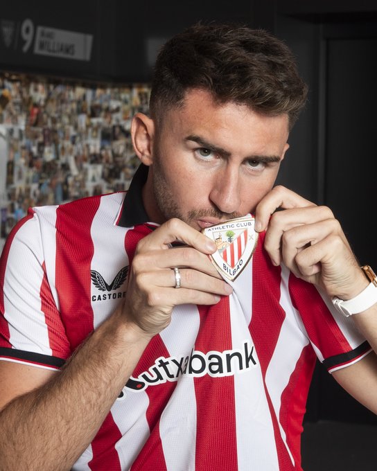 Aymeric Laporte wearing a red and white striped Athletic Club jersey with "Kutxabank" text on the front. He is holding and kissing a triangular badge featuring the Athletic Club logo. The background includes a wall with photos and the number 9 visible.