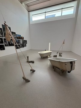 Two old bathtubs placed on the floor in a spacious, well-lit gallery room. Strings with high-heeled shoes and small sculptures of feet are suspended or positioned nearby. Photographs and framed images are mounted on the wall. Text on the image includes "MSN" and "Muzeum Sztuki Nowoczesnej w Warszawie" with additional details about the location and date.