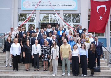 A group of people sitting around a large wooden table in a classroom, engaged in discussion. Shelves with books and educational materials line the walls. Children and adults planting a small tree in a garden, with some holding gardening tools. A large group of people, including adults and children, posing on the steps of a school building with a Turkish flag and a sign reading "Büyükada Şehit Murat Yüksel İlkokulu." Children sitting at tables in a classroom, working with crayons and papers, with an adult in a suit observing.