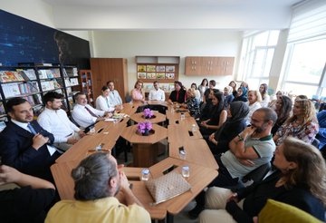 A group of people sitting around a large wooden table in a classroom, engaged in discussion. Shelves with books and educational materials line the walls. Children and adults planting a small tree in a garden, with some holding gardening tools. A large group of people, including adults and children, posing on the steps of a school building with a Turkish flag and a sign reading "Büyükada Şehit Murat Yüksel İlkokulu." Children sitting at tables in a classroom, working with crayons and papers, with an adult in a suit observing.