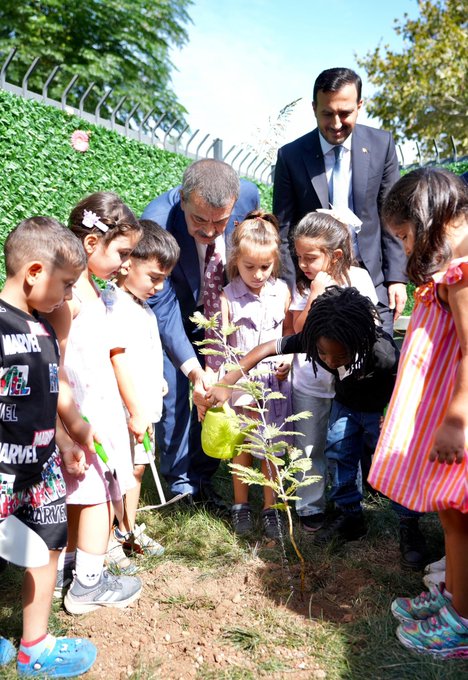 A group of people sitting around a large wooden table in a classroom, engaged in discussion. Shelves with books and educational materials line the walls. Children and adults planting a small tree in a garden, with some holding gardening tools. A large group of people, including adults and children, posing on the steps of a school building with a Turkish flag and a sign reading "Büyükada Şehit Murat Yüksel İlkokulu." Children sitting at tables in a classroom, working with crayons and papers, with an adult in a suit observing.