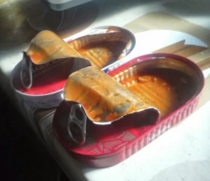 Two open sardine cans with red rims, filled with orange liquid, creatively arranged to resemble footwear. The cans have pull-tabs and are placed on a light-colored surface near a window, with sunlight casting shadows.