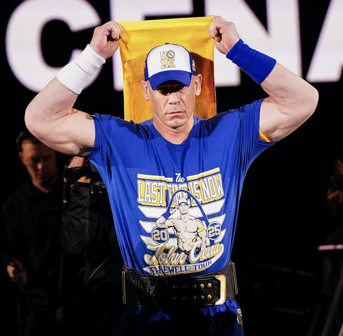John Cena wearing a blue T-shirt with "Last Time Ever 2025 John Cena Farewell Tour" text and an illustration, blue wristbands, a black belt, and a blue cap with a white "10" logo. He holds a yellow and blue flag above his head, displaying muscular arms.