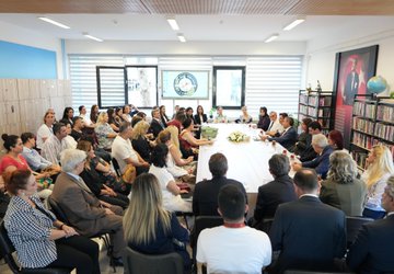 A group of people, including students in black uniforms and adults in formal attire, standing in rows on a yellow and green marked pavement outside a building with a sign reading "Halil Bedii Yönetken Ortaokulu." A man in a blue suit shakes hands with a student. Inside, people sit around a long table in a meeting room with bookshelves and a screen displaying a logo. Another image shows a large group of people, including students and adults, posing together on a red surface with banners in the background.