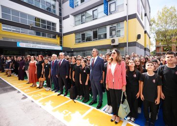 A group of people, including students in black uniforms and adults in formal attire, standing in rows on a yellow and green marked pavement outside a building with a sign reading "Halil Bedii Yönetken Ortaokulu." A man in a blue suit shakes hands with a student. Inside, people sit around a long table in a meeting room with bookshelves and a screen displaying a logo. Another image shows a large group of people, including students and adults, posing together on a red surface with banners in the background.