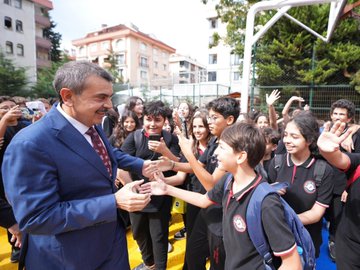 A group of people, including students in black uniforms and adults in formal attire, standing in rows on a yellow and green marked pavement outside a building with a sign reading "Halil Bedii Yönetken Ortaokulu." A man in a blue suit shakes hands with a student. Inside, people sit around a long table in a meeting room with bookshelves and a screen displaying a logo. Another image shows a large group of people, including students and adults, posing together on a red surface with banners in the background.