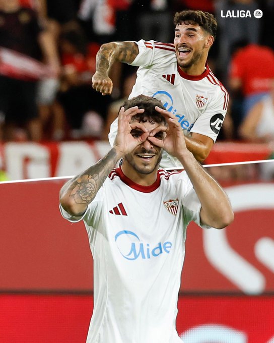 Two soccer players wearing white jerseys with red trim and the Mide logo, celebrating on a field. One player stands with arms raised, tattoos visible on his arms, while the other jumps behind him, making a hand gesture forming an "O" over his eyes. A red background with blurred spectators is visible, along with a LaLiga watermark in the top right corner.